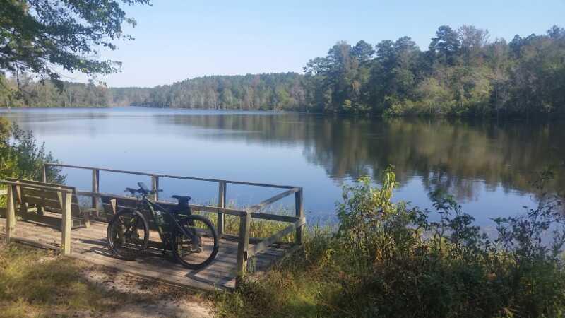 A serene lakeside scene featuring a wooden observation deck with a bicycle parked beside it. The calm water reflects the surrounding trees, creating a picturesque natural landscape under clear blue skies. Boyd Pond mountain bike trail.