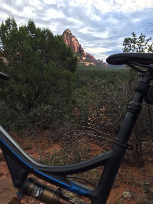 A mountain bike frame in the foreground, with a scenic view of red rock formations and green trees in the background, under a cloudy sky. Mescal Trail mountain bike trail.