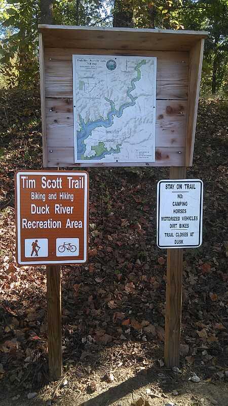 Signage at the Tim Scott Trail entrance, featuring a map of the Duck River Recreation Area. The left sign indicates it's for biking and hiking, while the right sign provides trail rules and restrictions, including no camping, horses, motorized vehicles, or dirt bikes, and notes that the trail closes at dusk. Surrounding environment shows autumn foliage. Duck River mountain bike trail.