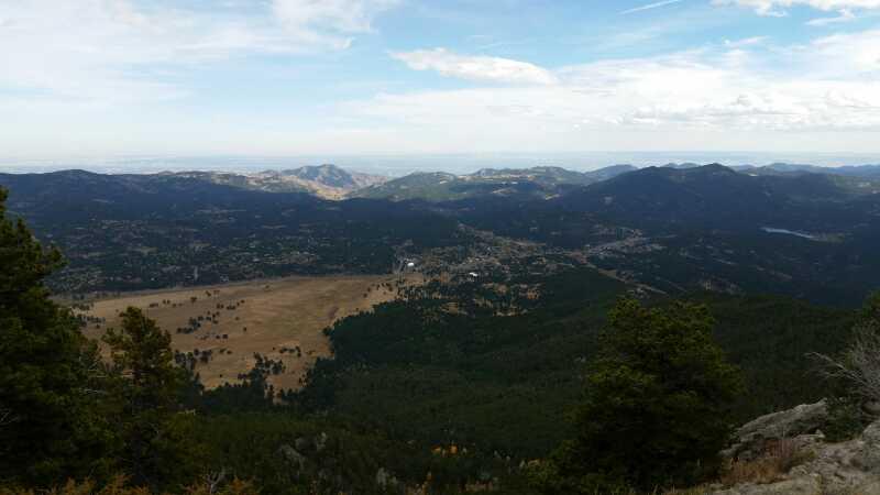 Panoramic view of a valley surrounded by mountains, featuring lush green forests and a grassy area in the foreground, under a partly cloudy sky. Elk Meadow mountain bike trail.