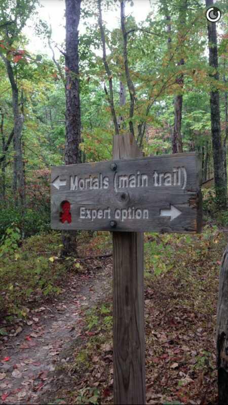 Wooden trail sign indicating two directions: "Mortals (main trail)" to the left and "Expert option" to the right, surrounded by trees and autumn foliage. The Trough mountain bike trail.