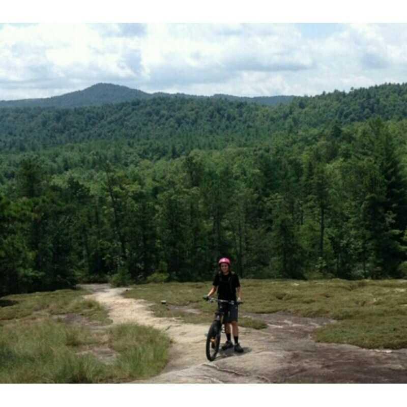 A person wearing a pink helmet stands beside a mountain bike on a rocky path, surrounded by lush green trees and rolling hills under a cloudy sky. Cedar Rock Trail #16 mountain bike trail.