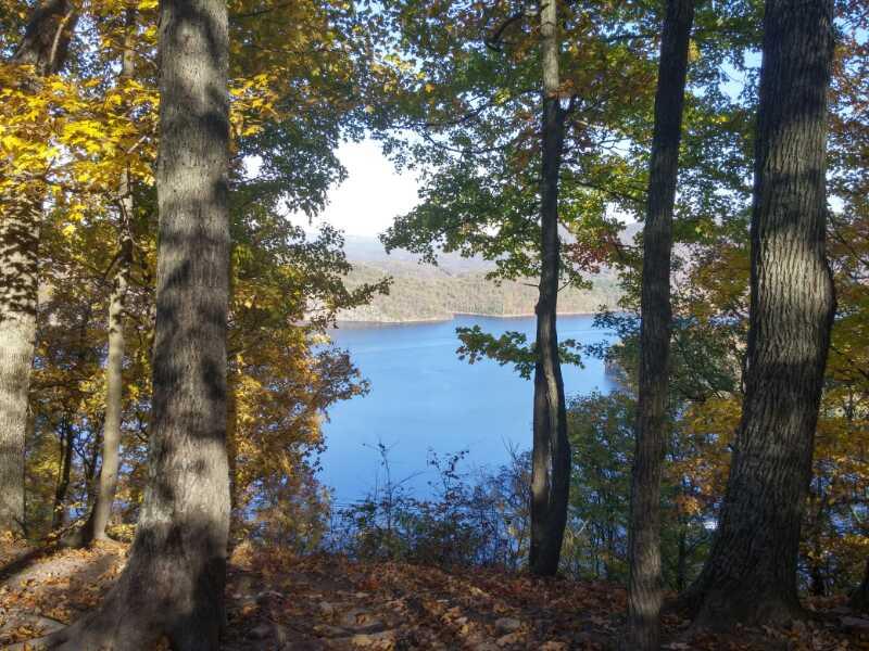 A serene view of a lake framed by trees with vibrant autumn foliage. The calm water reflects the clear blue sky and distant hills, creating a tranquil natural scene. Allegrippis Trails mountain bike trail.