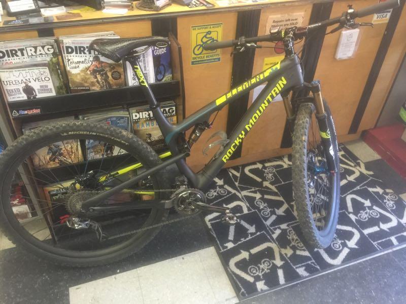 A Rocky Mountain mountain bike with a black and neon yellow design is positioned in a bike shop. The bike has a slightly muddy appearance, indicating recent use, and is placed on a patterned rug featuring bicycle silhouettes. Behind the bike, there are shelves filled with various cycling magazines and a sign that promotes bicycle repairs.