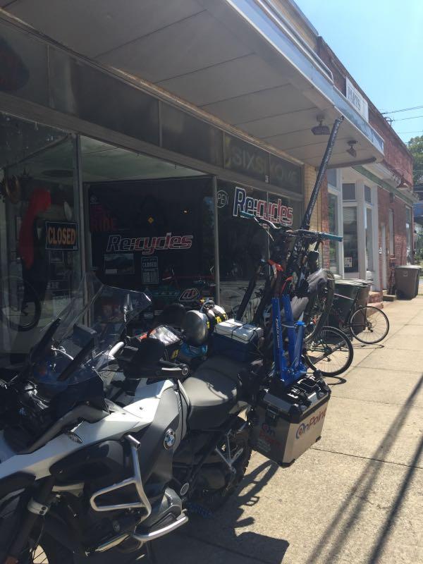 A storefront with large windows displaying a "CLOSED" sign. In the foreground, a parked motorcycle is partially visible, with various accessories and equipment attached to it. Next to the motorcycle, a bicycle is mounted on a blue rack. The shop's signage indicates it specializes in recycling and repair services. The setting appears to be a sunny day in an urban area.