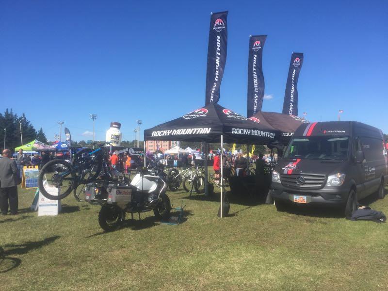 A lively outdoor event featuring a display area for Rocky Mountain bikes. There are several banners and a large tent showcasing bikes, with a van parked nearby. Attendees can be seen in the background engaging with various booths and exhibits under clear blue skies.
