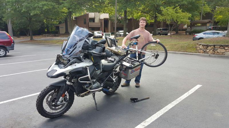 A person stands next to a motorcycle equipped with various accessories, including a mounted bicycle on the side. The motorcycle features a large windshield and is parked in a lot with trees and buildings in the background. The individual is smiling and appears to be adjusting or showcasing the attached bicycle.