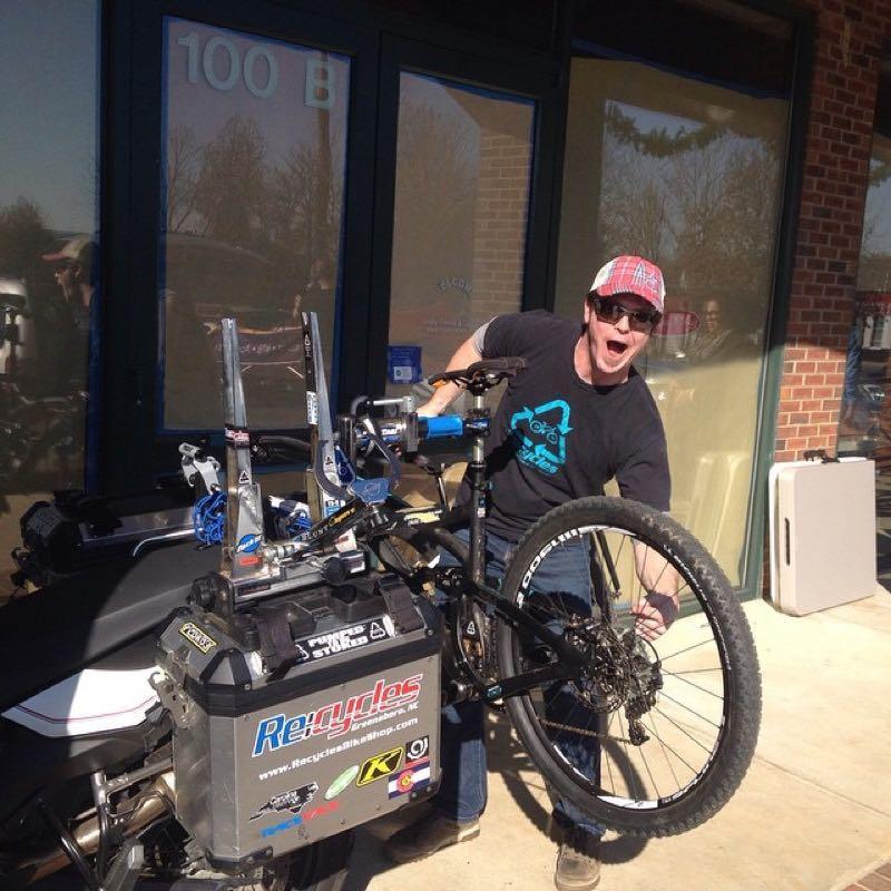 A person wearing a black t-shirt and a red baseball cap is enthusiastically inspecting a mountain bike while it is mounted on a bike rack. The bike has one wheel lifted off the ground, and various tools and gear are arranged nearby. A storefront with a sign reading "100 B" is visible in the background.
