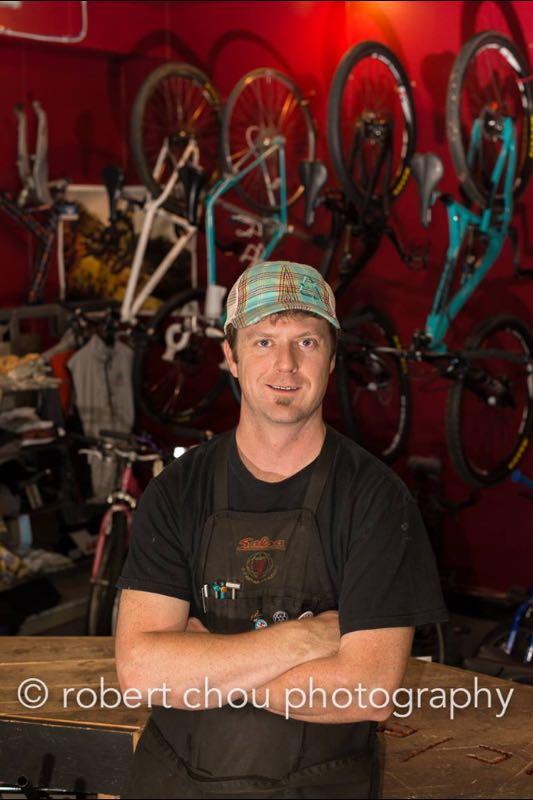 A bicycle mechanic stands with arms crossed in a bike shop, surrounded by various bicycles hanging on a vibrant red wall. He wears a black t-shirt and an apron with tools visible, showcasing a friendly and approachable demeanor.