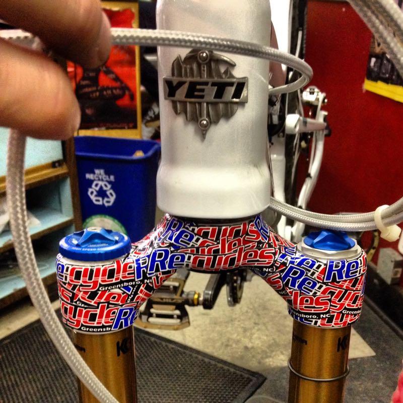 Close-up of a mountain bike's fork, featuring a silver Yeti logo at the top, decorated with colorful "Recycle" stickers in red and blue. The image shows one hand holding a cable, with the bike's suspension components visible below. A recycling bin sits in the background.