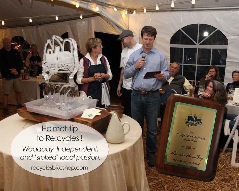 A festive indoor event under a tent, featuring people gathered around tables. In the foreground, a man is speaking into a microphone while holding a clipboard, presenting an award. An ice sculpture and a plaque recognizing "Re:cycles" as the 2015 Independent Business of the Year are visible. Guests in the background show expressions of interest and appreciation. Strings of lights hang overhead, creating a warm atmosphere.