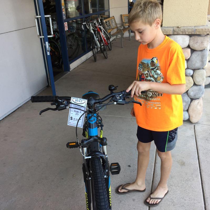 A young boy in an orange T-shirt and shorts is examining the handlebars of a blue bicycle outside a bike shop. The bike has a price tag attached, displaying a price of $376. In the background, other bicycles are visible on display.