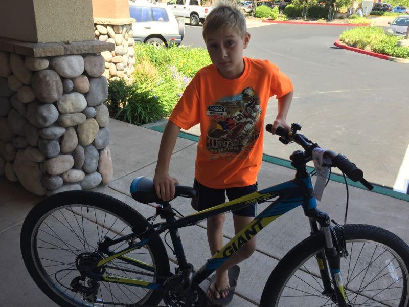A young boy wearing an orange t-shirt stands next to a blue and yellow mountain bike. He has short blonde hair and is wearing black shorts and flip-flops. The background features a stone wall and a green landscape with plants. The setting appears to be a parking area.