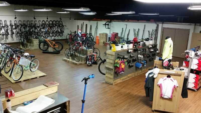 Interior of a bicycle shop featuring various bikes on display, sports apparel, and accessories. The shop has wooden displays and shelves showcasing helmets, clothing, and bike maintenance tools. The space is well-lit with a wooden floor, creating an inviting atmosphere for customers.