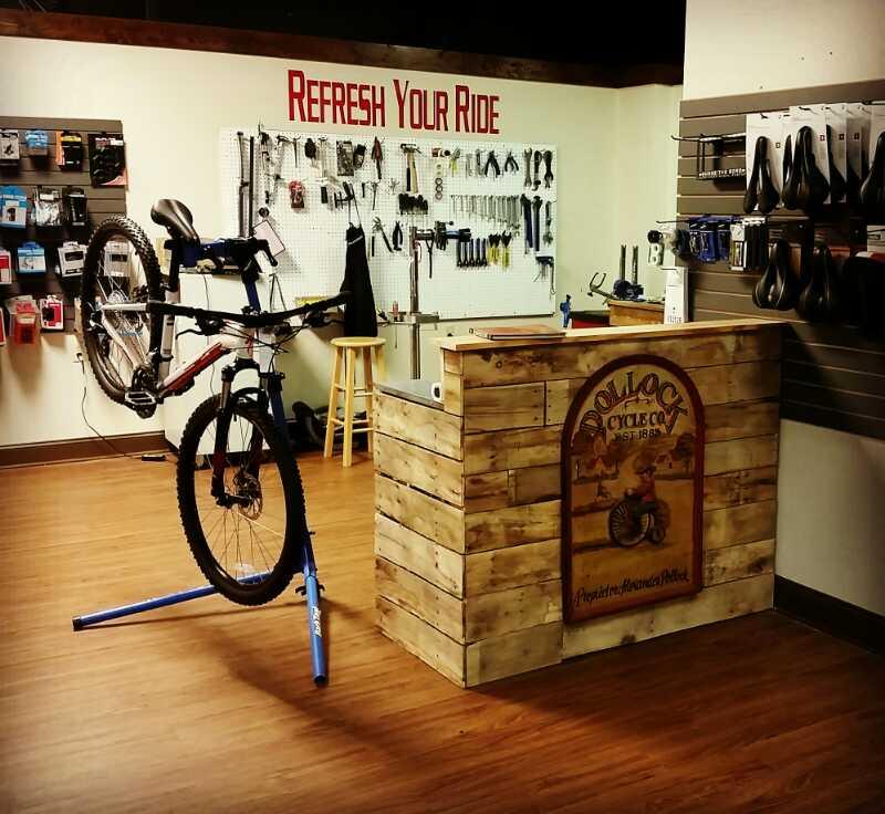 An interior view of a bicycle repair shop, featuring a bike on a repair stand, woodworking decor, and a service counter with signage. Various tools and accessories are displayed on the walls, with a motivational phrase "Refresh Your Ride" prominently above. The flooring is wooden, creating a welcoming atmosphere.