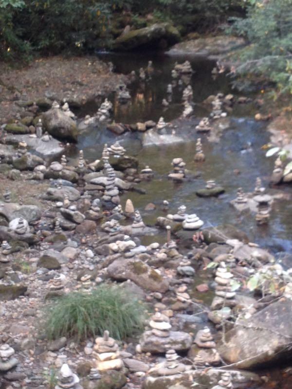 A serene stream surrounded by smooth stones and rocks, featuring numerous stacked stone cairns of varying heights scattered along the riverbanks. The lush green foliage in the background enhances the natural setting, creating a tranquil atmosphere. Black Mountain mountain bike trail.
