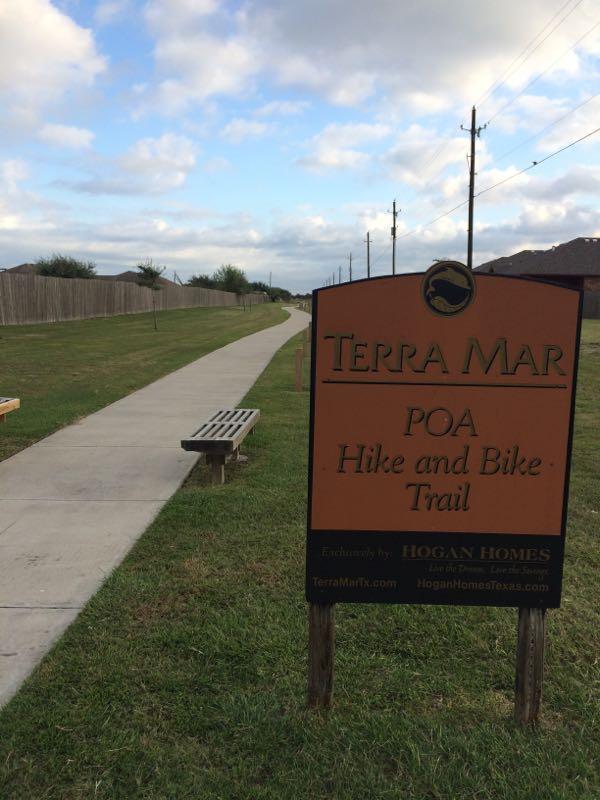 A sign indicating "Terra Mar POA Hike and Bike Trail" alongside a concrete path. The area features green grass, benches for resting, and power poles in the background under a cloudy sky. Holly Hike and Bike trails mountain bike trail.