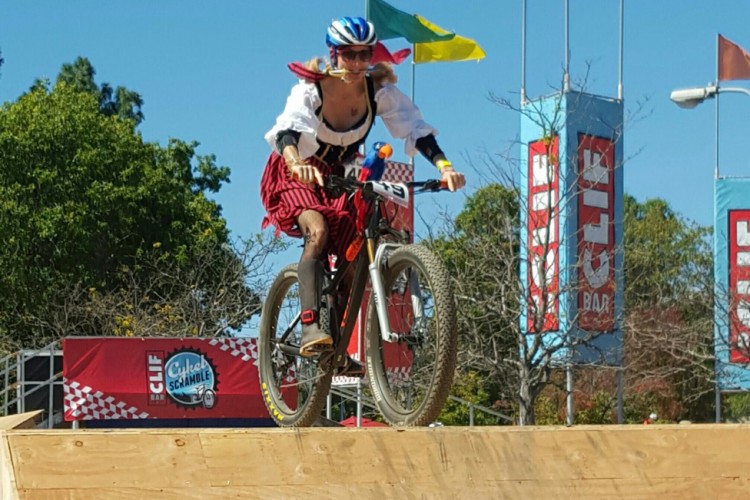 A person wearing a colorful costume, featuring a white top and a red skirt, is riding a mountain bike down a wooden ramp at an outdoor event. The background includes flags and banners, indicating a festive atmosphere. The sky is clear and blue, enhancing the vibrant scene.