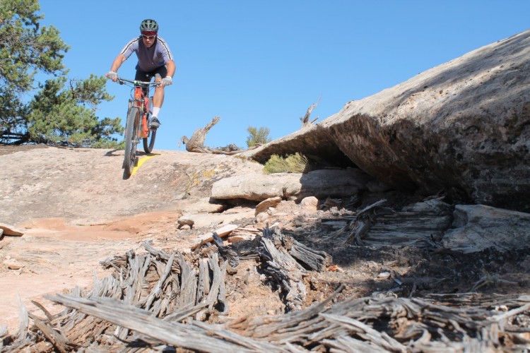 A person riding a mountain bike over rocky terrain, navigating a challenging landscape under a clear blue sky. The cyclist is focused on the trail ahead, with trees and natural features in the background.