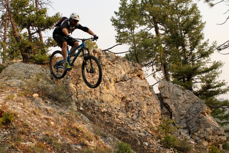 A mountain biker in mid-air jumps off a rocky ledge, surrounded by trees and a natural landscape, showcasing an adventurous outdoor scene.