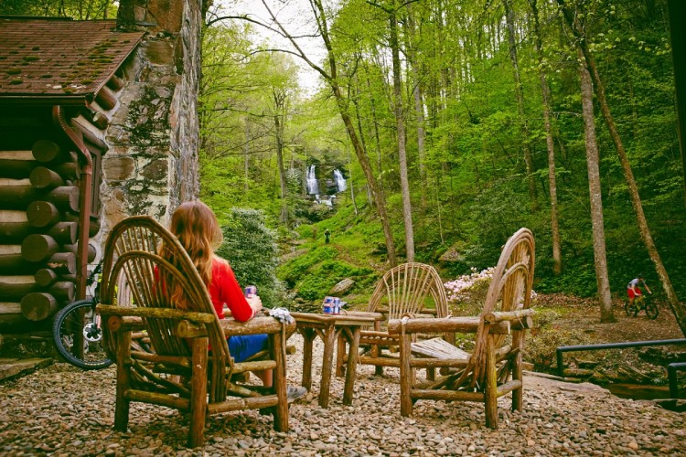 A woman sitting in a rustic wooden chair, enjoying a drink while overlooking a forested landscape with a waterfall in the background. In the scene, there are additional wooden chairs arranged around a small table, and a mountain biker can be seen on a trail nearby, surrounded by greenery and blooming flowers.