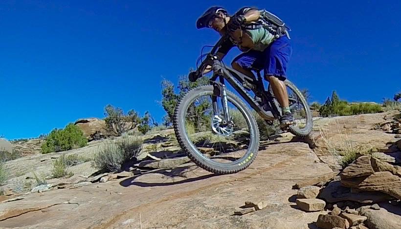 A mountain biker in mid-jump over rocky terrain, showcasing an action-packed moment on a sunny day with a clear blue sky. Surrounding vegetation includes sparse shrubs and trees, highlighting an outdoor adventure setting. Killer Bee mountain bike trail.