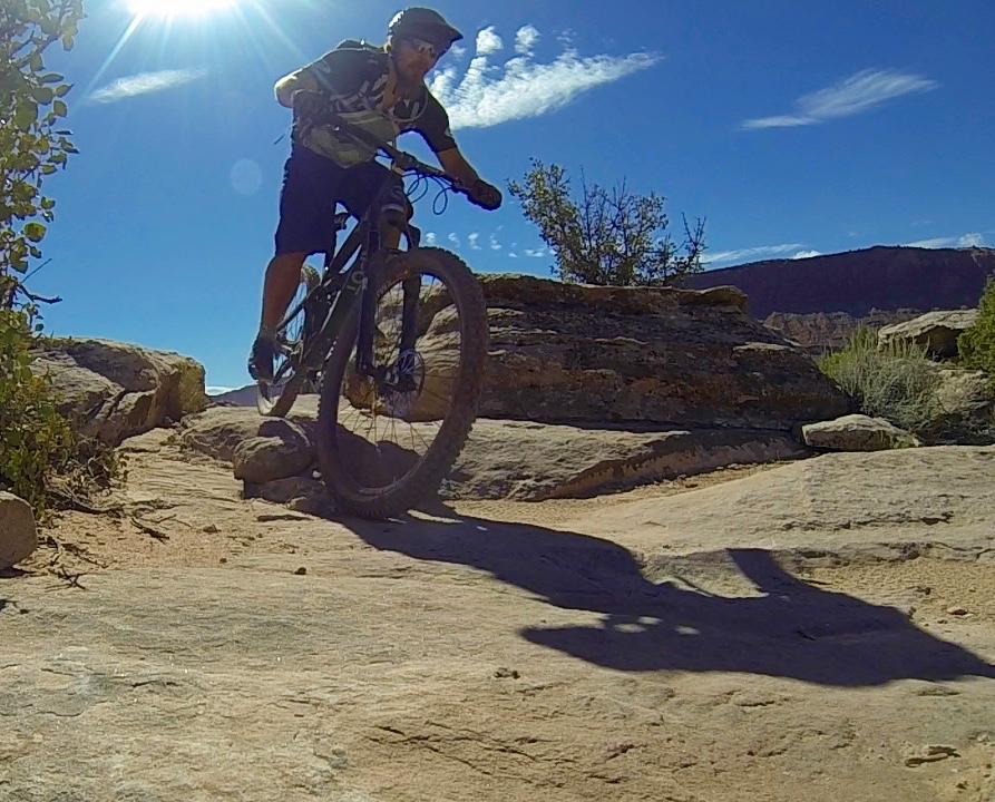 A mountain biker navigating rocky terrain under a bright sun, with blue skies and wispy clouds in the background. The biker is in motion, showcasing skillful maneuvering on a rugged path. Vegetation and rock formations are visible on either side of the trail. Moab Brand Trails mountain bike trail.
