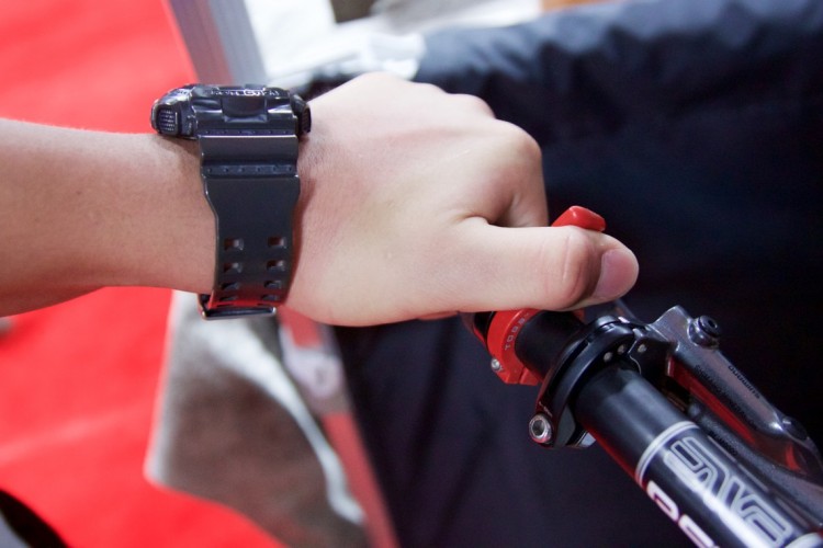 A close-up image of a hand gripping a red trigger lever on a mountain bike handlebar, wearing a black sports watch on the wrist. The background features a blurred red carpet.