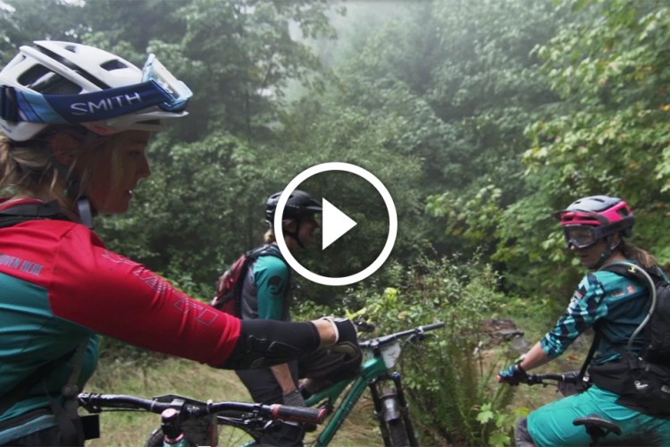 Three mountain bikers in colorful gear are gathered in a lush, green forest, discussing their ride. The environment is foggy, enhancing the adventurous atmosphere. Two of the bikers are wearing helmets and goggles, while one is gesturing towards the trail. Mountain bikes are positioned nearby, indicating a pause in their ride.