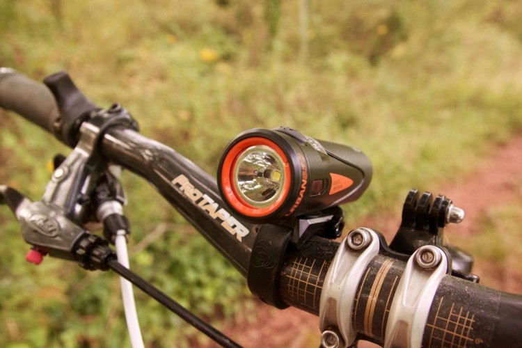 Close-up of a bicycle handlebar featuring a mounted headlight, set against a blurred natural background. The handlebar has a "ProTaper" logo and visible brake levers, with the headlight designed for outdoor use.