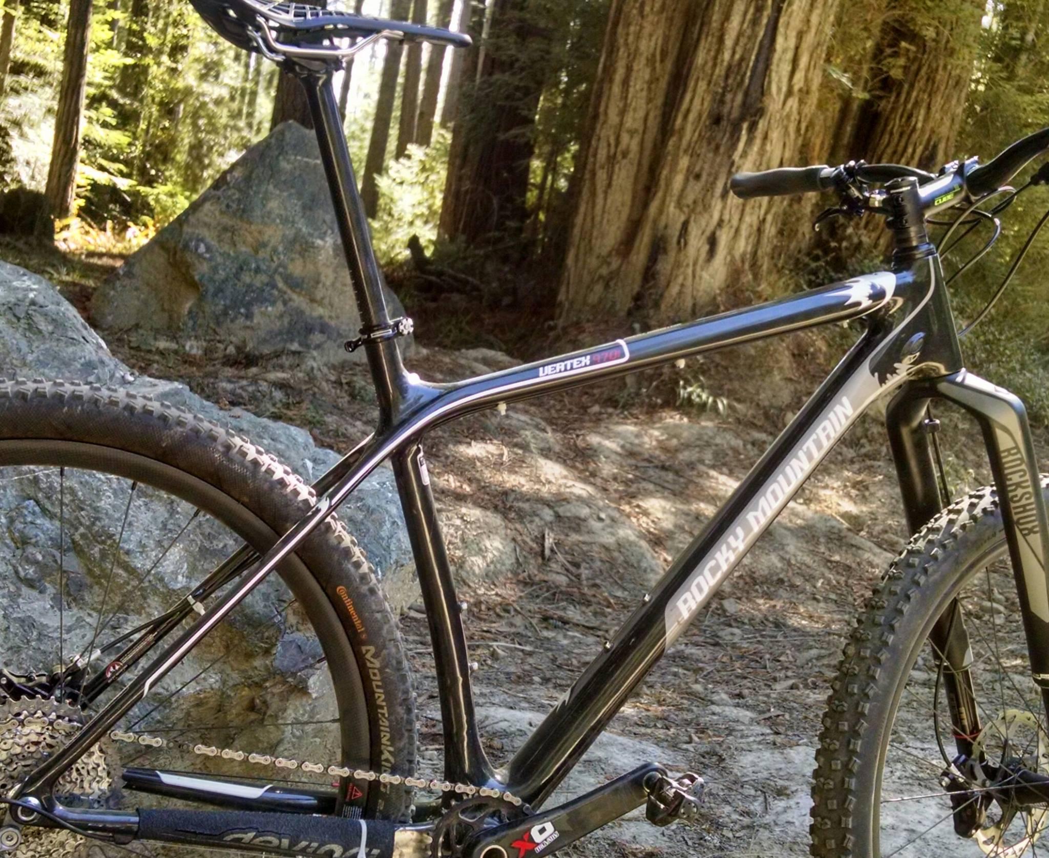 Rocky Mountain Vertex: A close-up view of a black mountain bike, featuring its frame, handlebars, and wheel. The bike is positioned on rocky terrain, surrounded by tall trees in a natural outdoor setting. The frame displays the brand name "Rocky Mountain," and the background includes a mix of greenery and large stones.