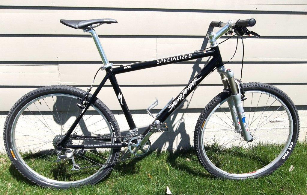 Specialized Stumpjumper Comp: A black Specialized Stumpjumper mountain bike positioned upright on green grass, with a background of light-colored wooden siding. The bike features distinctive frame geometry, front suspension forks, and knobby tires suitable for off-road riding.