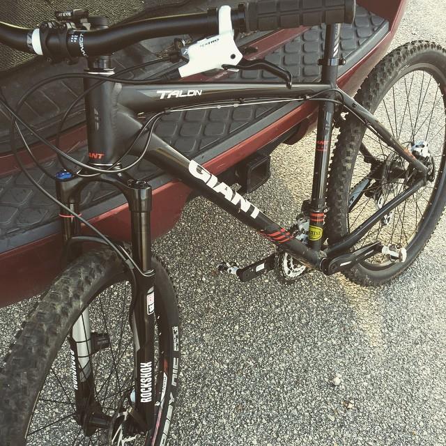Giant Talon 27.5: A black mountain bike with the brand name "GIANT" and model "Talon" is positioned on a textured surface, partially visible against the backdrop of a vehicle. The bike features front suspension and knobby tires, indicating its suitability for off-road use.