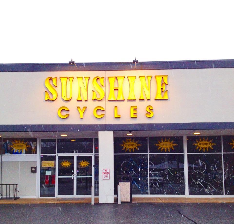 Front view of a bike shop named "Sunshine Cycles," featuring large yellow lettering on a storefront. The store has glass windows displaying bicycles inside, and there are signs in the window. The scene is set in snowy weather.
