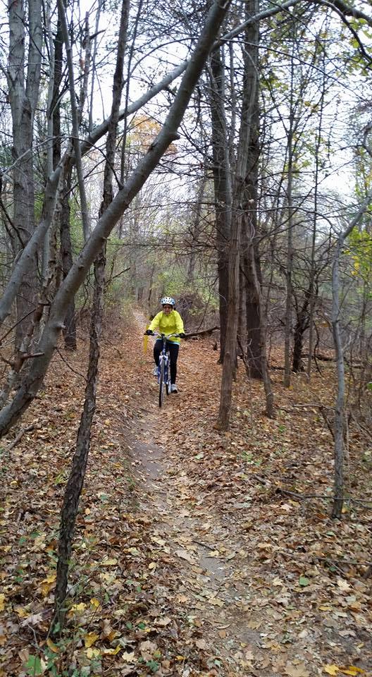 A cyclist wearing a bright yellow jacket rides a mountain bike down a dirt path surrounded by trees in a forest during autumn. The ground is covered with fallen leaves, and the atmosphere is overcast. Maybury mountain bike trail.
