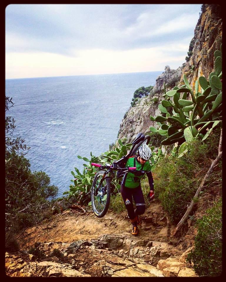 A mountain biker carrying their bike up a rocky trail alongside a cliff that overlooks the ocean. The person is wearing a green and black cycling outfit, a helmet, and is navigating a steep, rugged path surrounded by vegetation and cacti. The sky is overcast, adding to the adventurous atmosphere of the scene. bikeridecostabrava mountain bike trail.