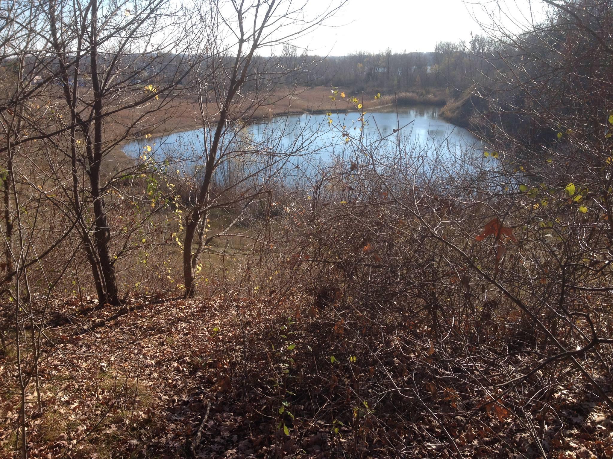 A tranquil scene of a small, reflective lake surrounded by bare trees and sparse foliage. The area is covered with dry leaves, indicating a late autumn or early winter setting. The water is calm, with a hint of blue sky visible above, creating a peaceful nature landscape. Pontiac Lake mountain bike trail.