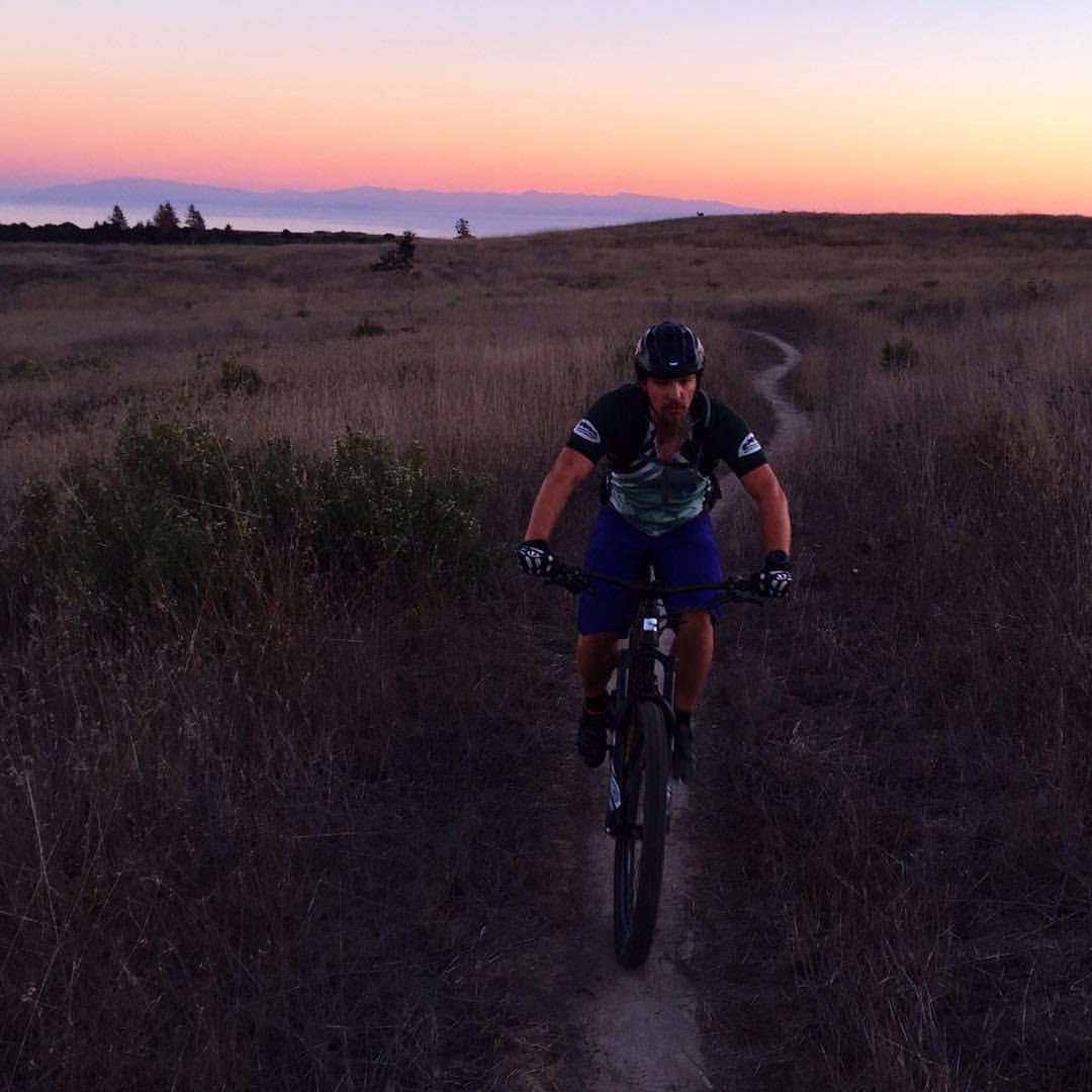 A mountain biker riding on a narrow dirt trail through tall grass, set against a colorful sunset sky with soft hues of pink and orange. The landscape features distant hills and trees silhouetted against the horizon. Wilder Ranch State Park mountain bike trail.