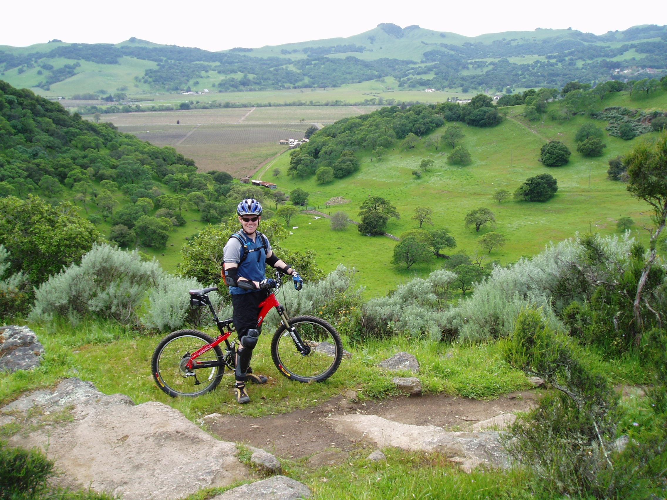 Jamis Dakar XLT: A person in biking gear stands next to a red mountain bike on a grassy hillside, overlooking a lush green valley with trees and farmland in the background. The landscape features rolling hills under a cloudy sky.
