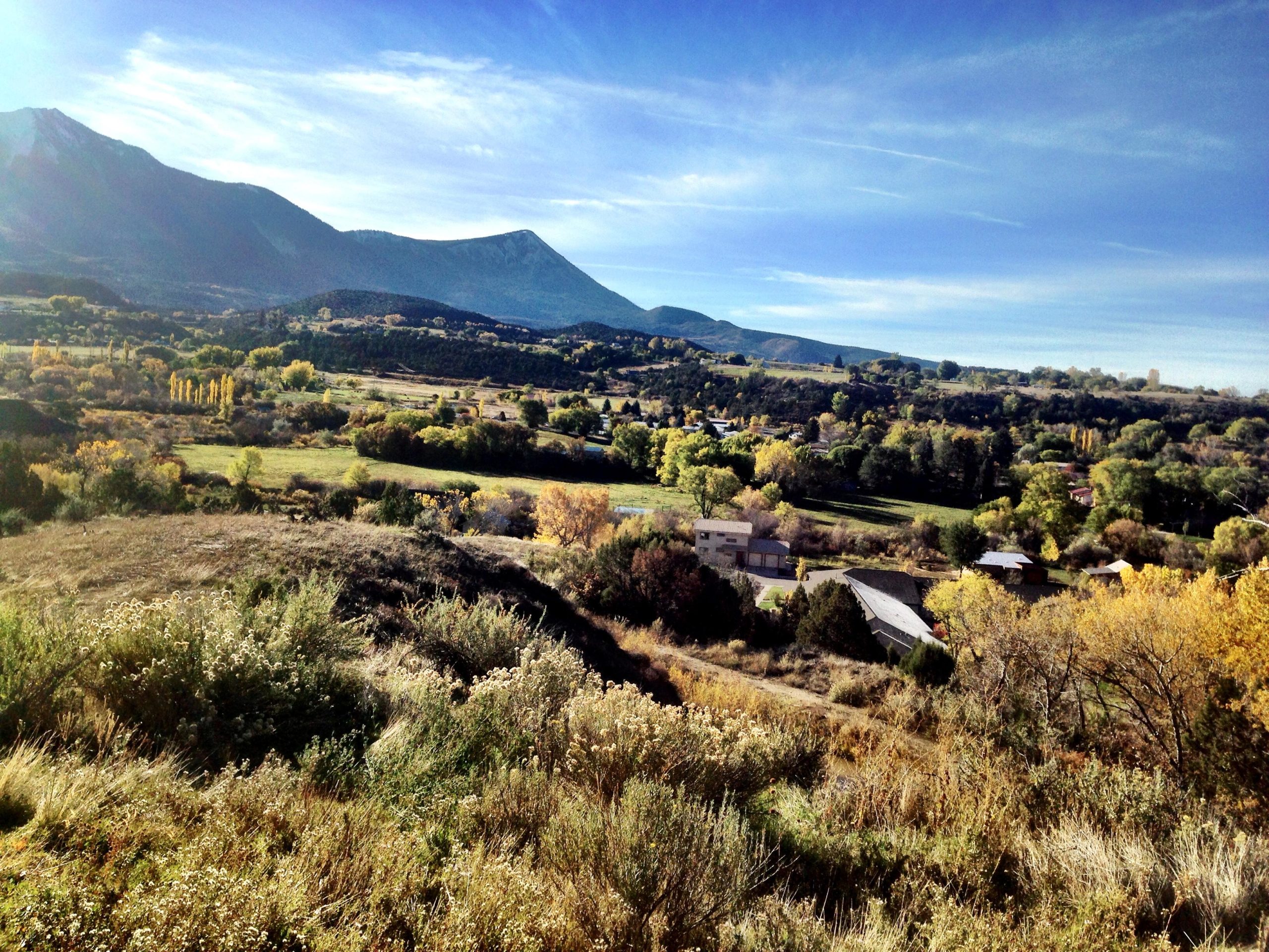 A picturesque landscape showcasing rolling hills and mountains under a clear blue sky. The foreground features grassy slopes dotted with wildflowers, while the background reveals a valley filled with trees displaying autumn colors. A small community of houses can be seen nestled among the greenery, with mountains rising majestically in the distance. Jumbo Mountain mountain bike trail.