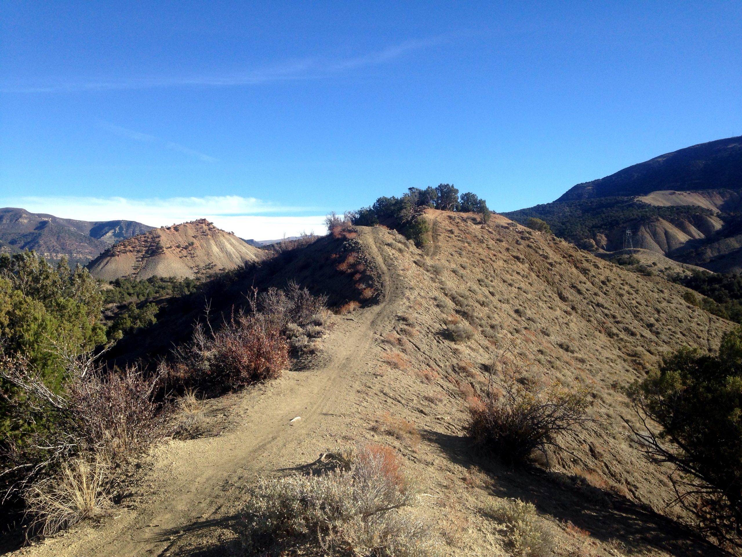 A winding dirt path leads through a rugged landscape featuring rocky hills and sparse vegetation under a clear blue sky. The scene showcases layered earth formations and distant mountains, creating a serene outdoor environment. Jumbo Mountain mountain bike trail.