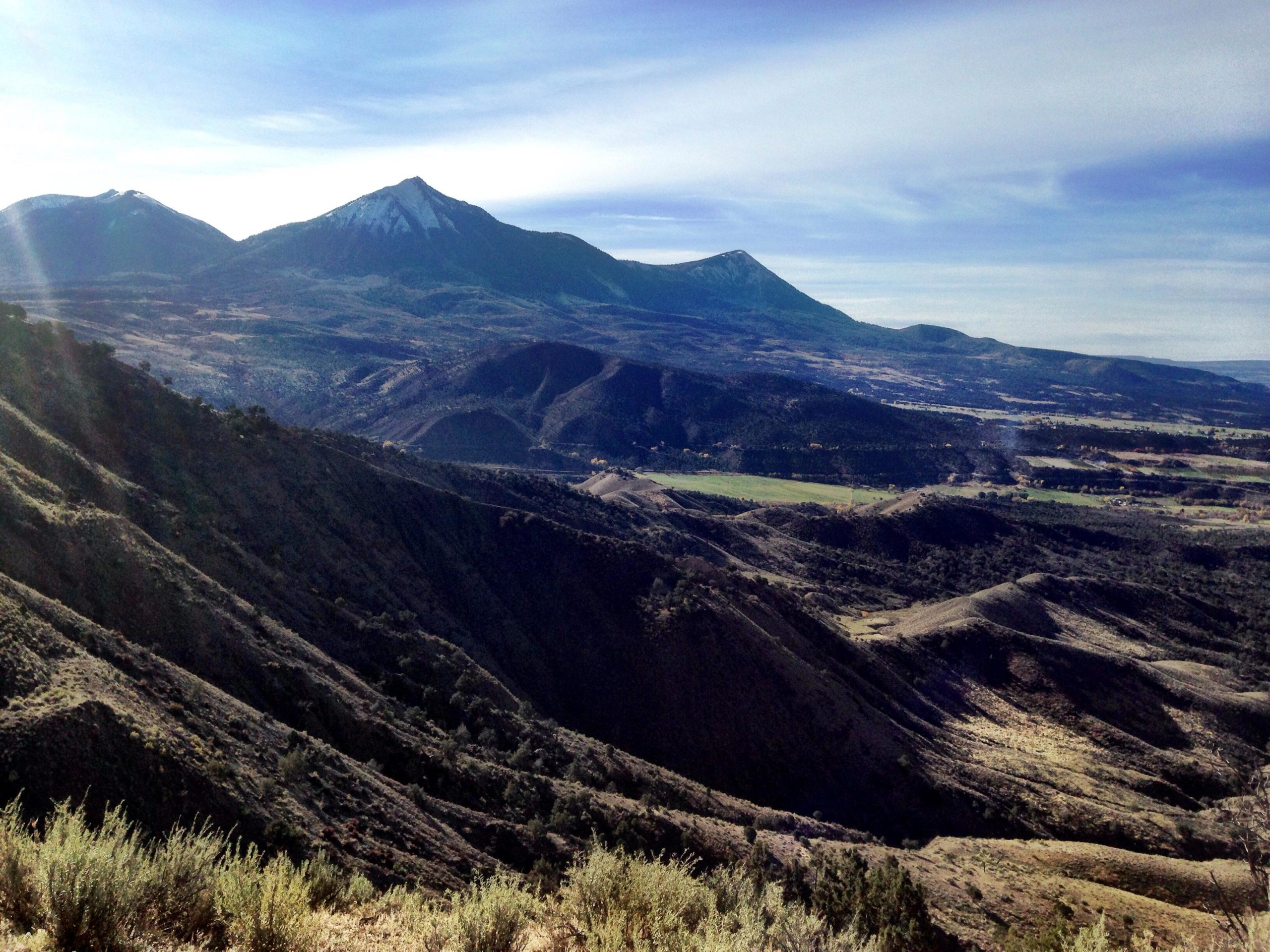 A panoramic view of a mountainous landscape featuring multiple peaks, some capped with snow, surrounded by rugged terrain and rolling hills. The sky is clear with soft clouds, and patches of green vegetation can be seen in the valley below. Jumbo Mountain mountain bike trail.