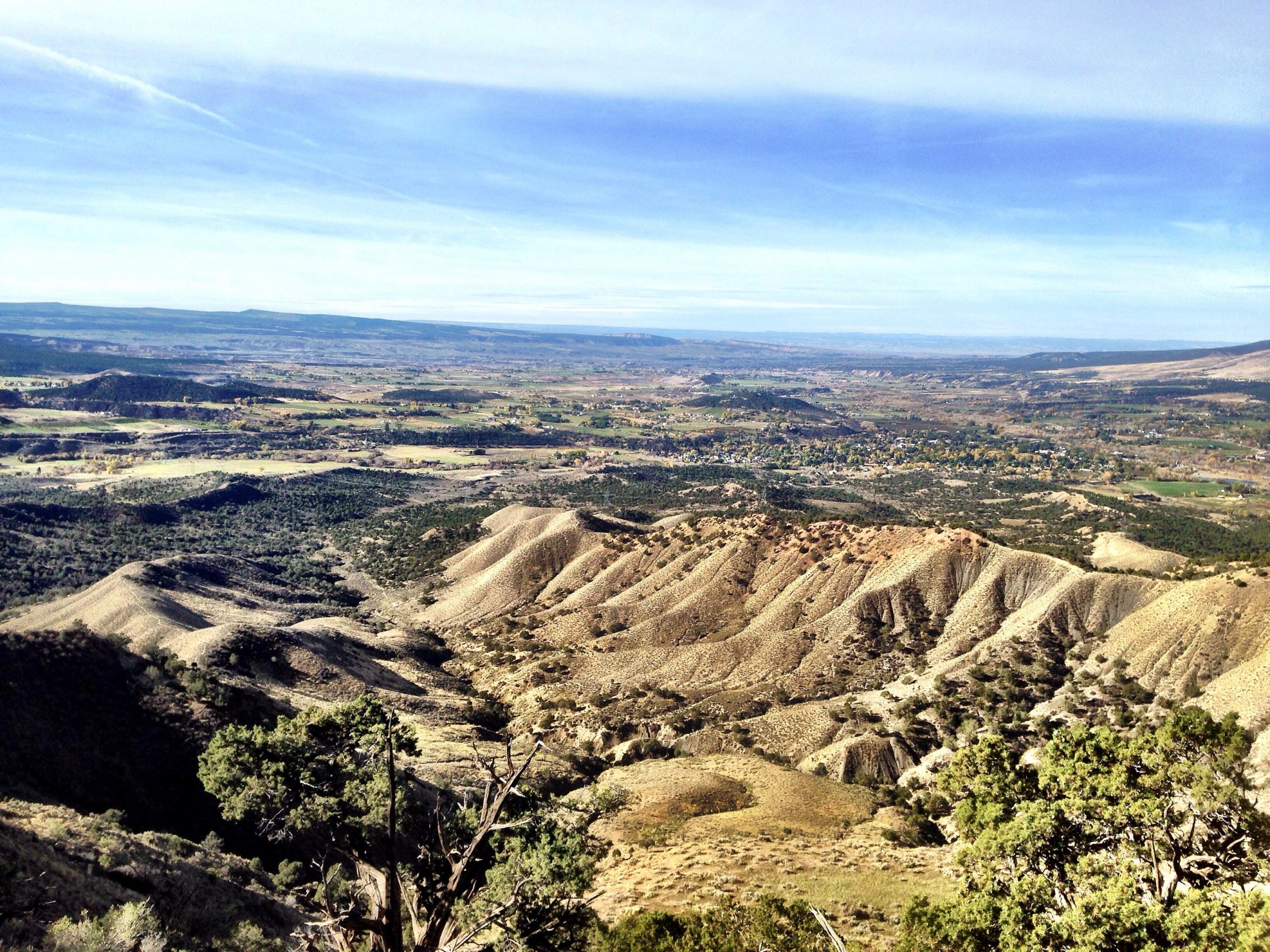 A panoramic view of a rugged landscape featuring rolling hills and valleys under a clear blue sky. The foreground showcases textured, hilly terrain with patches of greenery, while the distance reveals a blend of farmland and natural vegetation. Jumbo Mountain mountain bike trail.
