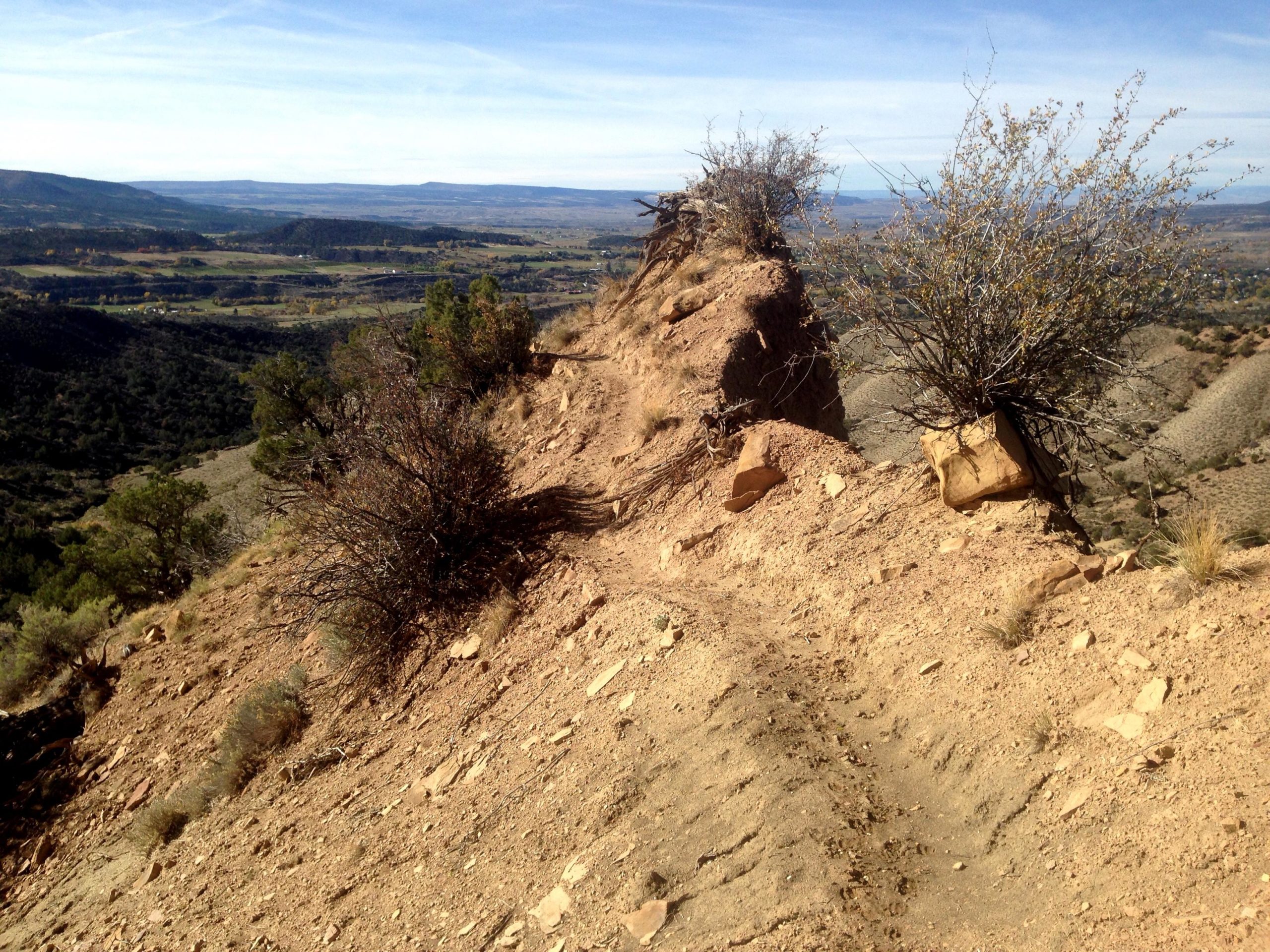 A rocky, dirt trail leading along a ridge with sparse vegetation, overlooking a vast valley filled with shrubs and hills under a clear blue sky. Jumbo Mountain mountain bike trail.