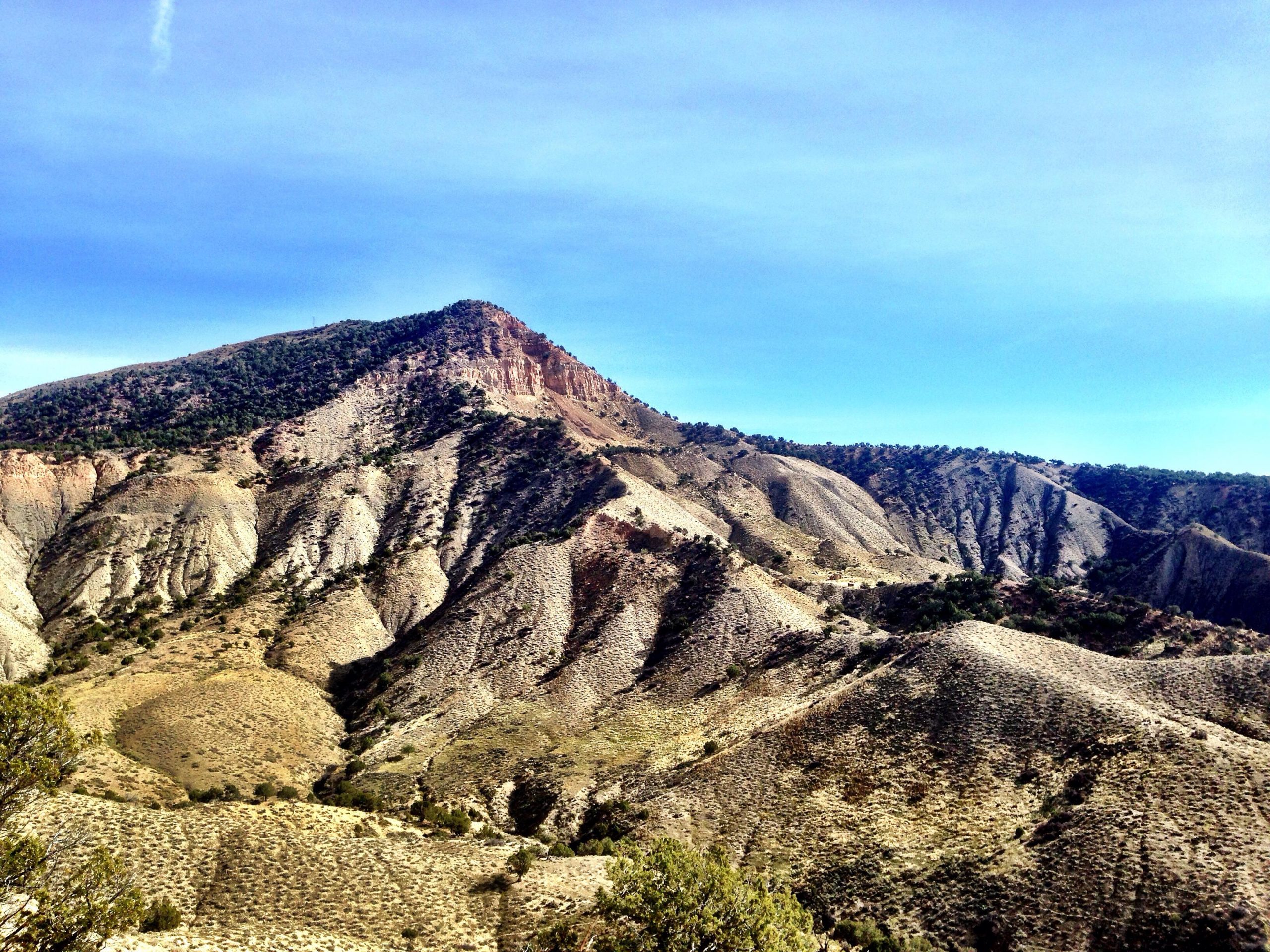 A panoramic view of a mountainous landscape featuring rugged terrain with pronounced slopes and ridges. The scene is characterized by a mix of earthy tones and patches of greenery, under a clear blue sky. The prominent peak in the background is surrounded by rolling hills and valleys, creating a sense of depth and natural beauty. Jumbo Mountain mountain bike trail.