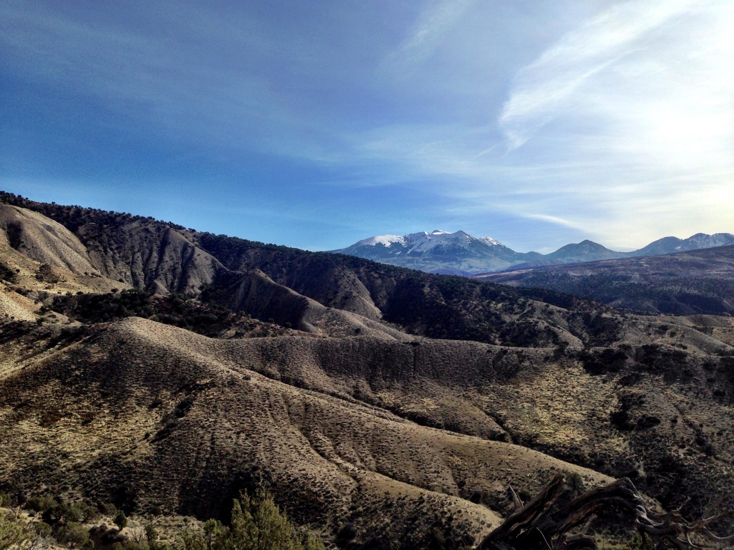 A panoramic view of rugged mountainous terrain under a clear blue sky, featuring rolling hills and distant snow-capped peaks. The landscape is dotted with sparse vegetation, showcasing natural earth tones. Jumbo Mountain mountain bike trail.