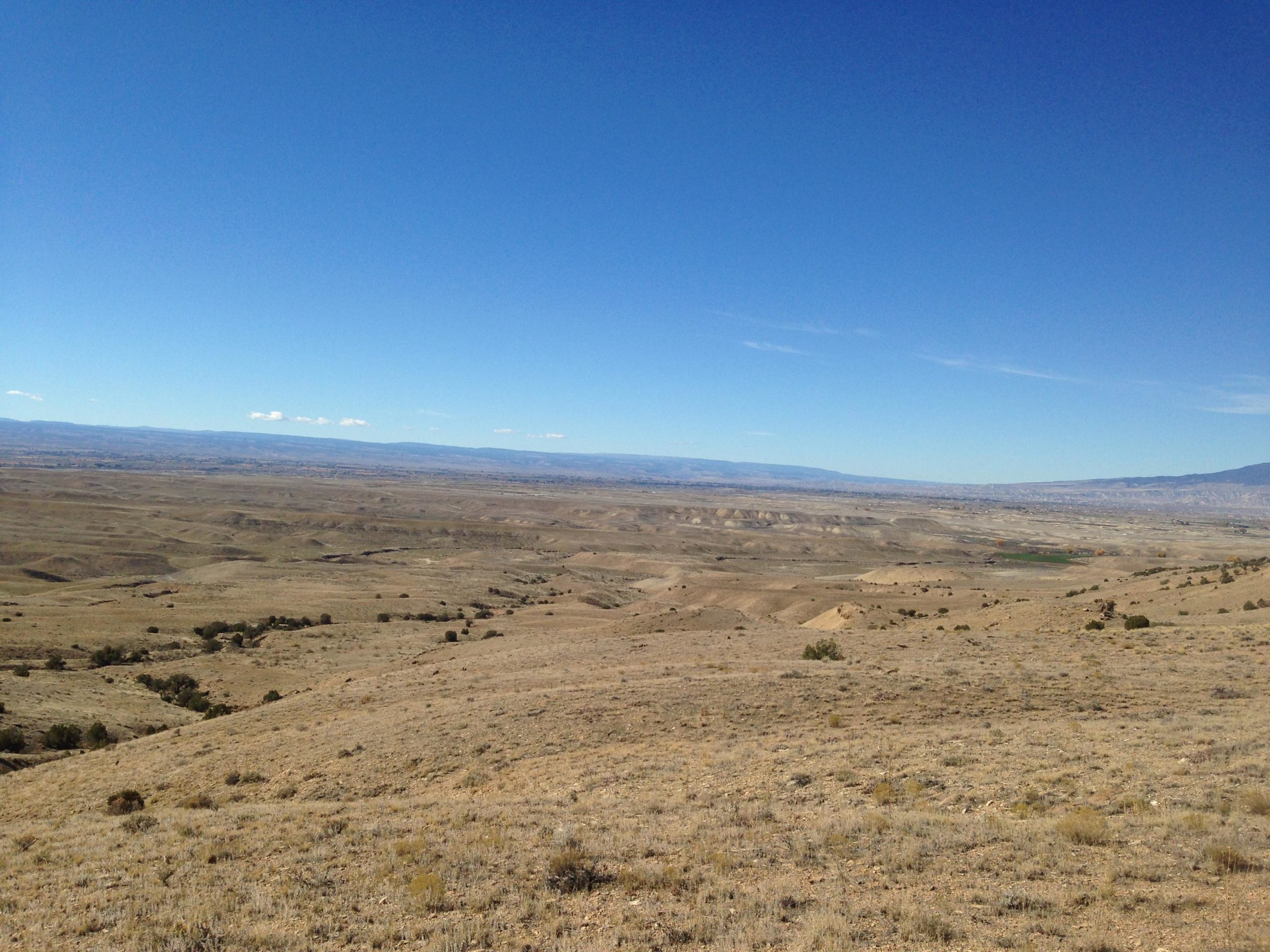 A panoramic view of an arid landscape, featuring rolling hills and valleys under a clear blue sky. The terrain is mostly barren with patches of greenery, showcasing natural variations in color and texture across the dry ground. Sidewinder mountain bike trail.
