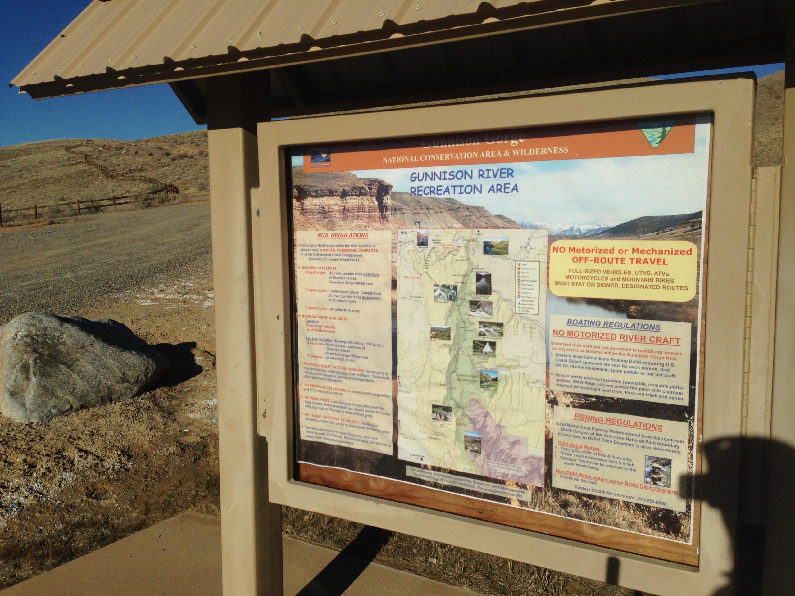 Information board for the Gunnison River Recreation Area, featuring regulations for visitors, including boating and fishing guidelines, alongside a map of the area. The sign is situated outdoors with a gravel path and a rocky landscape in the background. Sidewinder mountain bike trail.