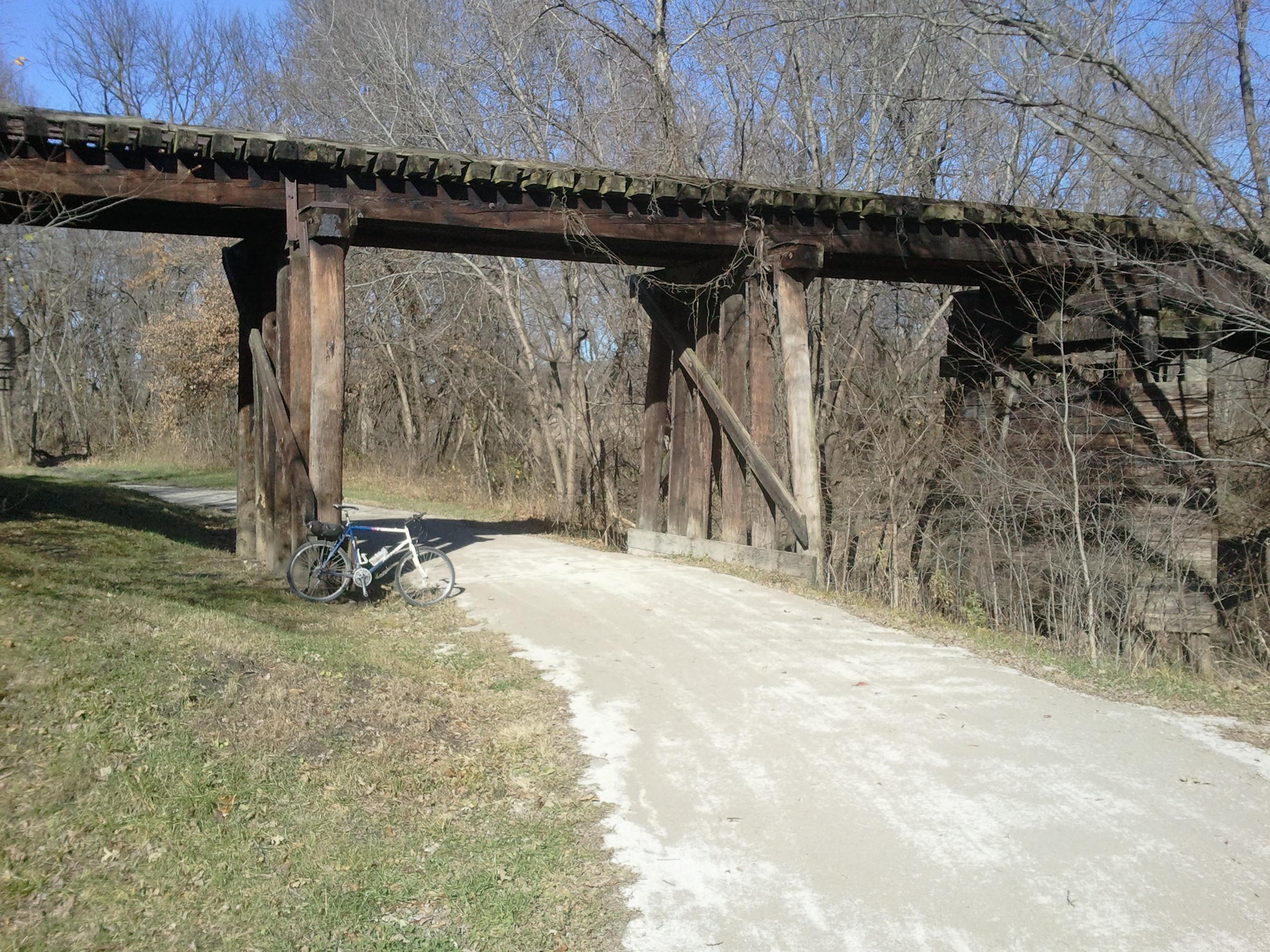 A white bicycle rests on a dirt path beside a weathered wooden bridge, surrounded by bare trees and grassy areas. The sky is clear and blue, creating a serene outdoor scene. Little Blue Trace mountain bike trail.