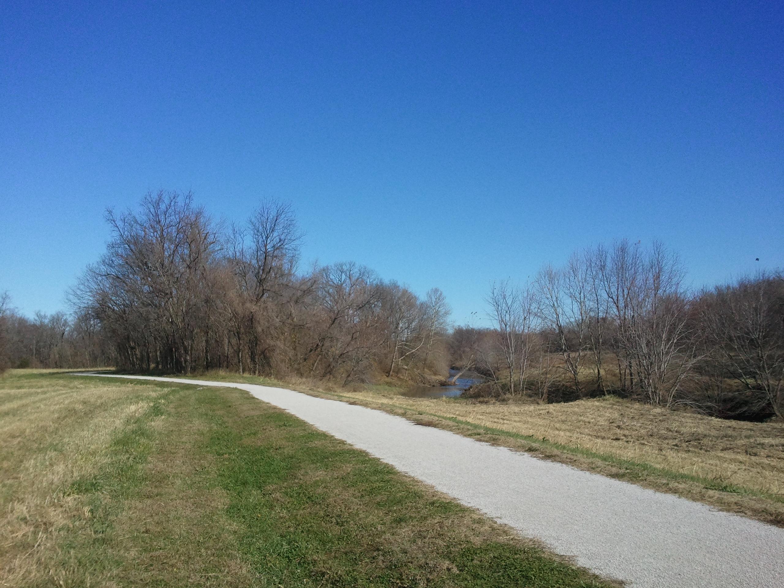 A peaceful outdoor scene featuring a winding gravel path surrounded by bare trees and grassy areas under a clear blue sky. A small stream runs parallel to the path. Little Blue Trace mountain bike trail.